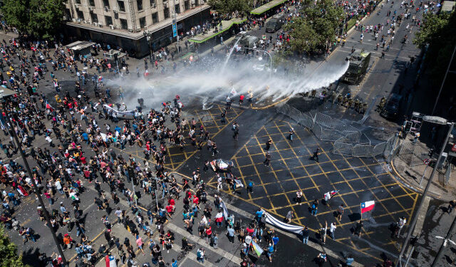 Aerial view showing riot police spraying water at demonstrators in Santiago, on the sixth straight day of street violence which erupted over a now suspended hike in metro ticket prices, on October 23, 2019. - A four-year-old child was killed during the latest round of protests against economic inequality in Chile, raising the death toll from five days of social unrest to 18 as unions launched a general strike on Wednesday. (Photo by Javier TORRES / AFP)