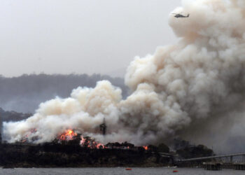 A military helicopter flies above a burning woodchip mill in Eden, in Australia's New South Wales state on January 6, 2020. - January 5 brought milder conditions, including some rainfall in New South Wales and neighbouring Victoria state, but some communities were still under threat from out-of-control blazes, particularly in and around the town of Eden in New South Wales near the Victorian border. (Photo by SAEED KHAN / AFP)