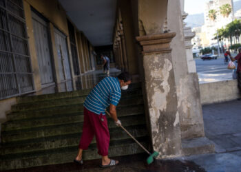 A man wears a face mask and gloves as a precautionary measure against the spread of the new coronavirus, COVID-19, while sweeping the sidewalk with soap water outside at the entrance of a  residential building, while  in Caracas on March 26, 2020. - Venezuela is facing the novel coronavirus pandemic while suffering a major gasoline shortage and with the country's water system collapsed, which has left many homes without running water. (Photo by Cristian Hernandez / AFP)