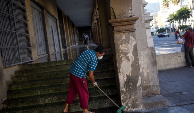 A man wears a face mask and gloves as a precautionary measure against the spread of the new coronavirus, COVID-19, while sweeping the sidewalk with soap water outside at the entrance of a  residential building, while  in Caracas on March 26, 2020. - Venezuela is facing the novel coronavirus pandemic while suffering a major gasoline shortage and with the country's water system collapsed, which has left many homes without running water. (Photo by Cristian Hernandez / AFP)