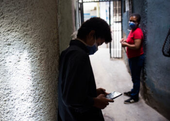 Jhonatan Figueroa, 14, uses his cellphone, connected to a neighbour's wifi signal, outside his home, as his mother Viviana Rodriguez looks at him, at the Bello Campo neighborhood in Chacao, Caracas, on October 6, 2020, amid the new coronavirus pandemic. - With students that have to catch internet in the streets and teachers who earn 2,5 US dollars a month, classes start up again in Venezuela. (Photo by Cristian Hernandez / AFP)