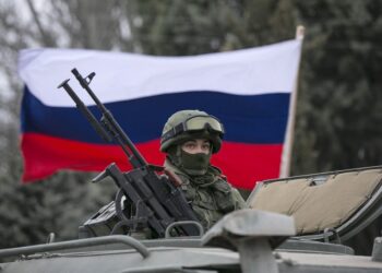 A pro-Russian man (not seen) holds a Russian flag behind an armed servicemen on top of a Russian army vehicle outside a Ukrainian border guard post in the Crimean town of Balaclava March 1, 2014. Ukraine accused Russia on Saturday of sending thousands of extra troops to Crimea and placed its military in the area on high alert as the Black Sea peninsula appeared to slip beyond Kiev's control. REUTERS/Baz Ratner (UKRAINE - Tags: MILITARY POLITICS CIVIL UNREST) - RTR3FVD5
